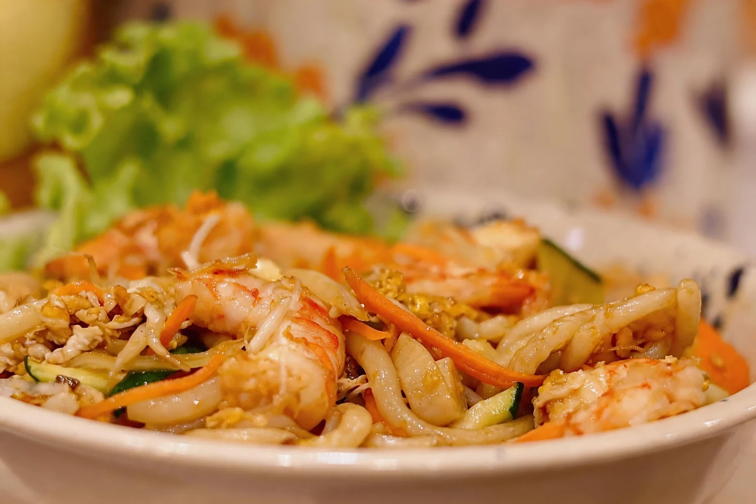 Close-up of stir-fried noodles with shrimp, julienned carrots and green salad in the background.